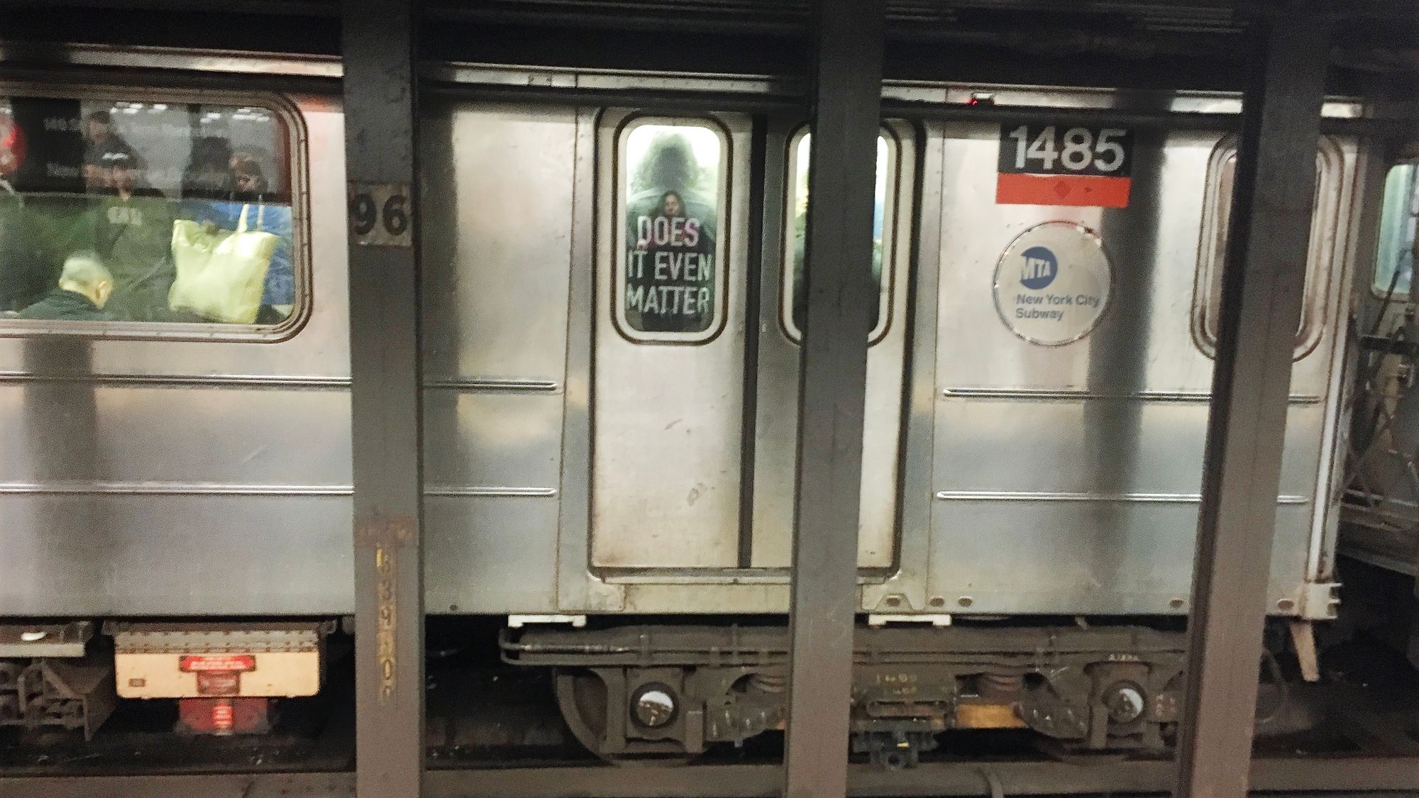 The text on the back of a MTA's passengers jacket reads "DOES IT EVEN MATTER", visible through a train door.